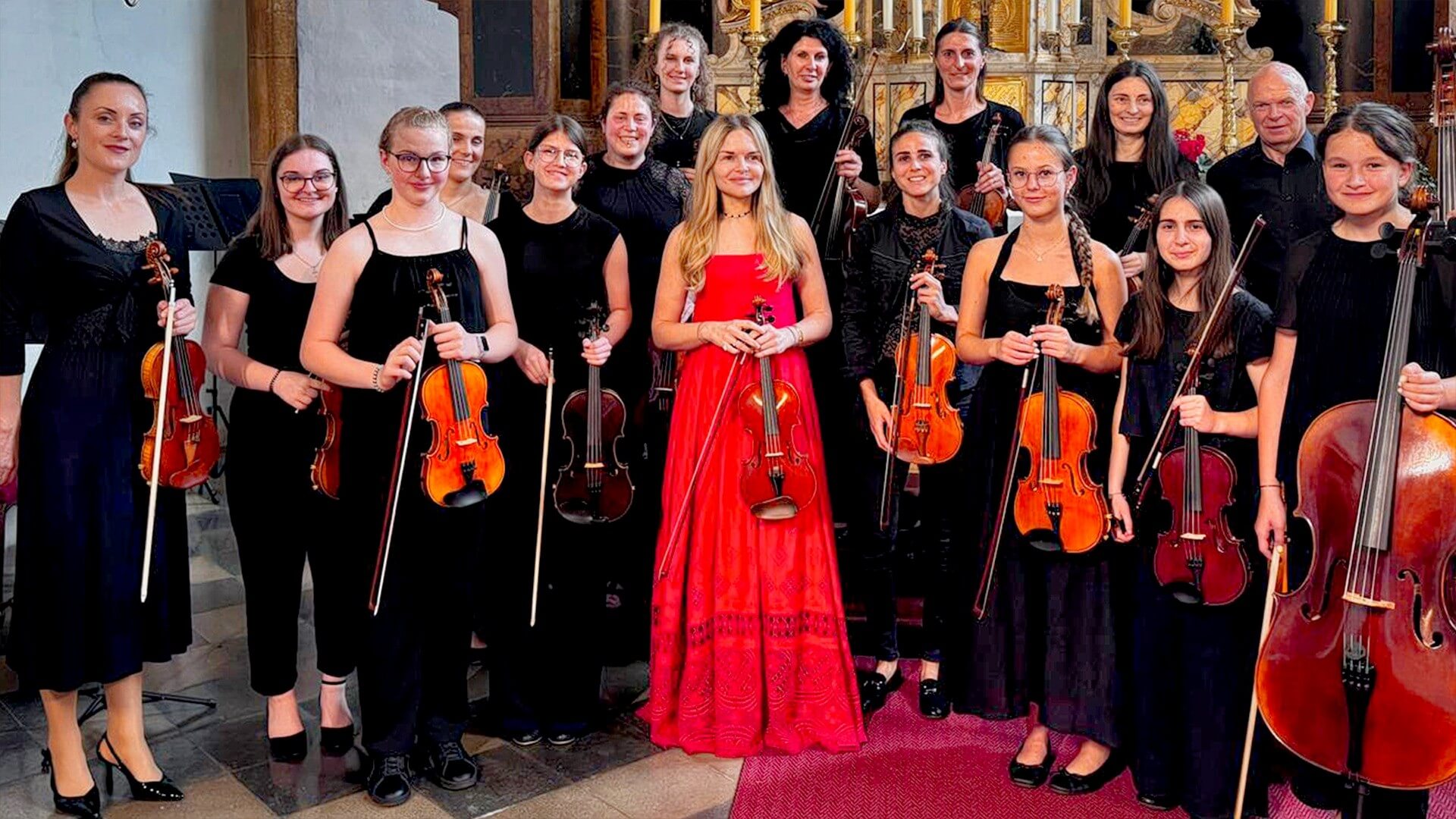Eine Gruppe von Frauen und Mädchen in Schwarz posiert mit Geigen, während eine Frau in einem roten Kleid im Vordergrund steht.