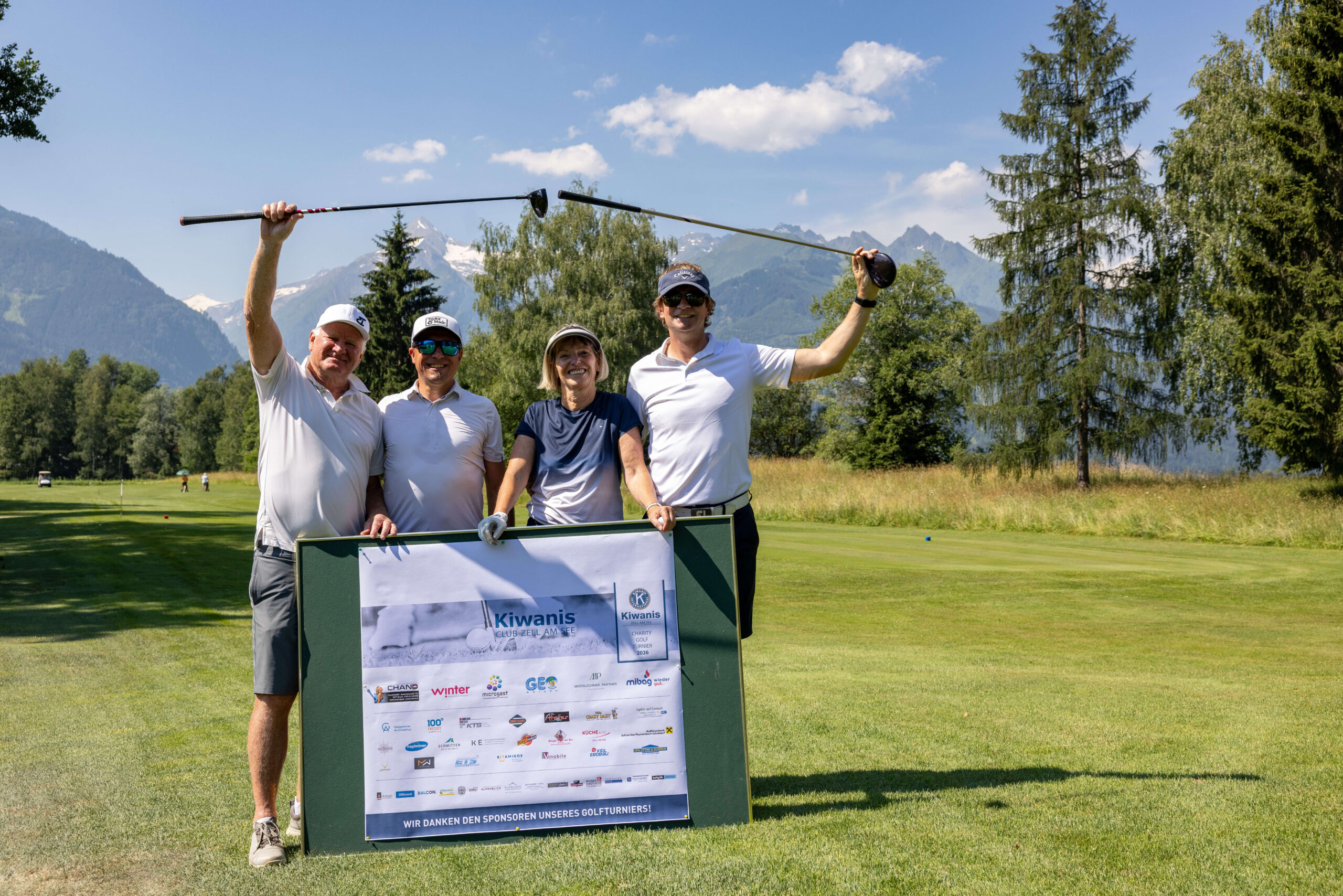 Vier Golfer stehen lächelnd vor einem Schild mit Sponsorenlogos, umgeben von einer malerischen Landschaft und Bergen.