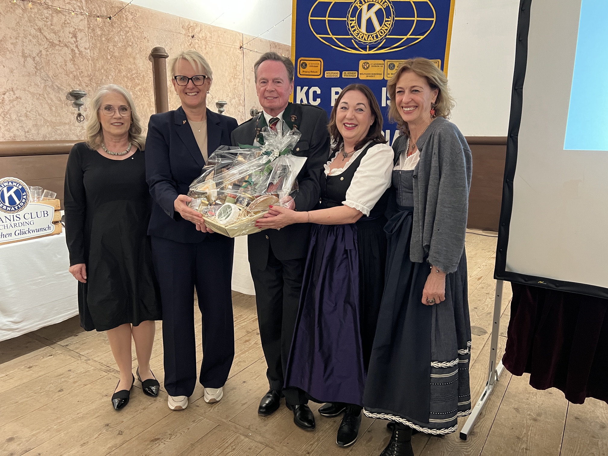 Fünf Personen stehen zusammen, eine überreicht einen Geschenkkorb, während im Hintergrund Dekoration und ein Präsentationsbild zu sehen sind.