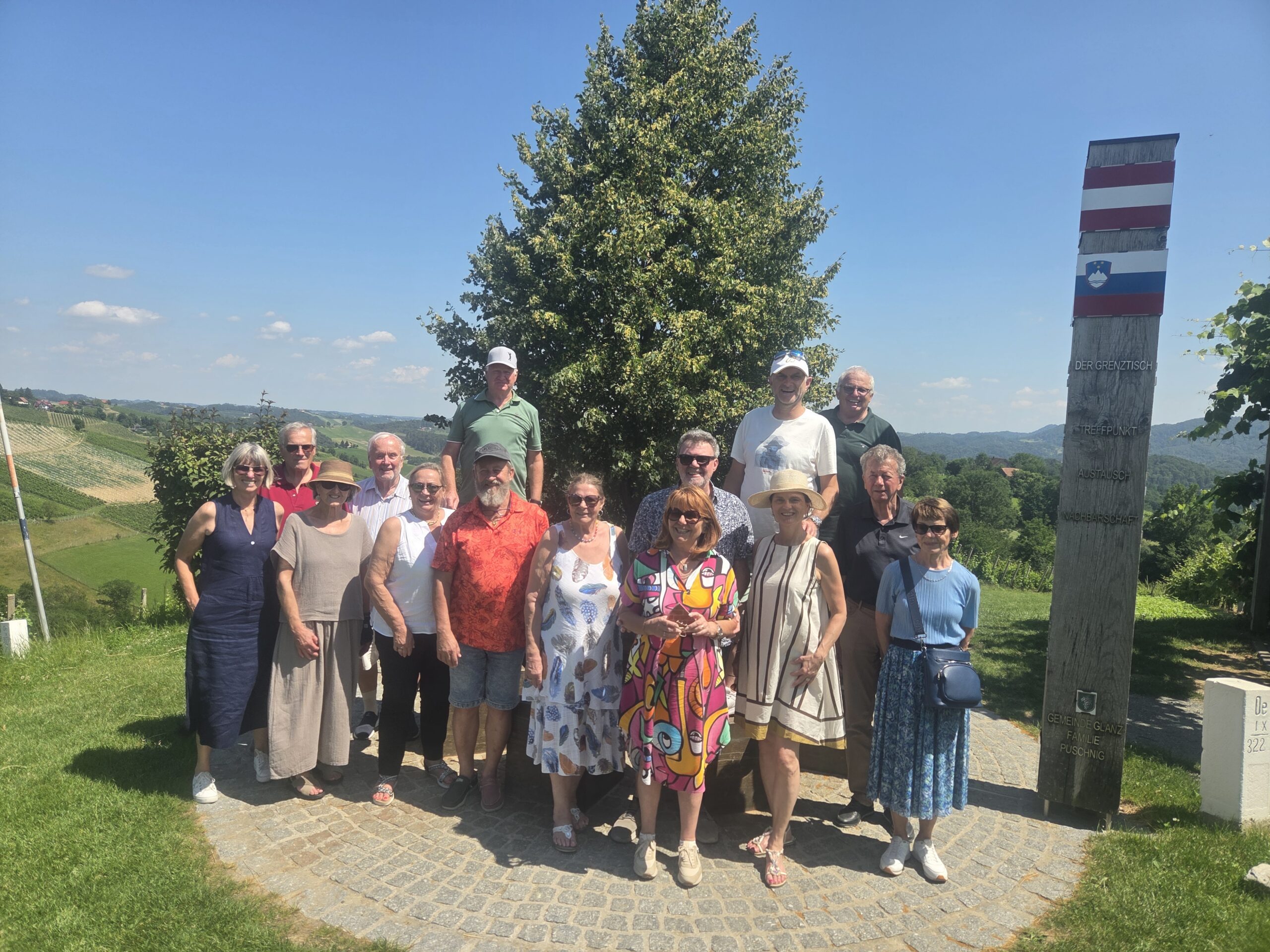 Gruppe von 18 Personen in heller Kleidung steht vor einem Baum und einer Grenzsteinmarkierung bei sonnigem Wetter.