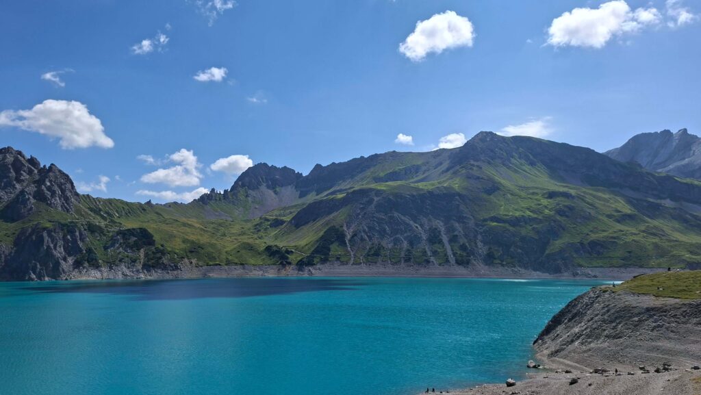 Blick auf einen klaren, türkisfarbenen See umgeben von grünen Bergen und einem blauen Himmel mit einigen Wolken.