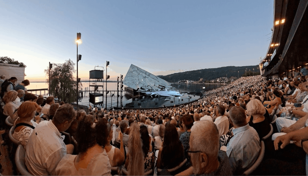 Eine große Menschenmenge sitzt in einem Freilufttheater mit einer modernen Bühne am Wasser während des Sonnenuntergangs.