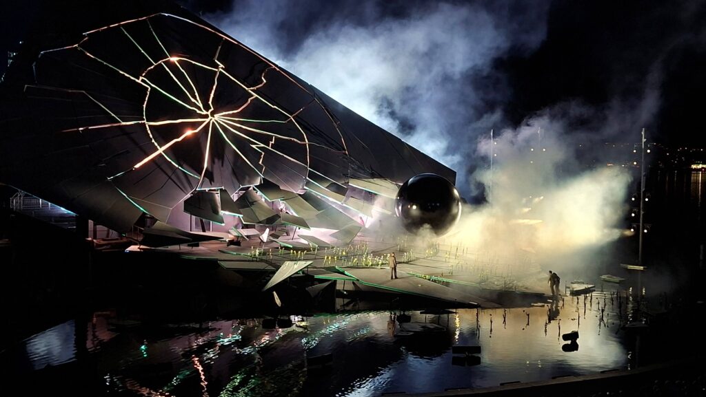 Eine futuristische Bühne mit zerbrochenen Elemente, einem großen schwarzen Ball und Nebel, reflektiert im Wasser.