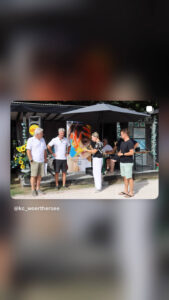 Vier Personen stehen im Freien vor einem Pavillon, während eine Frau mit einem Mikrofon spricht und ein buntes Plakat präsentiert.