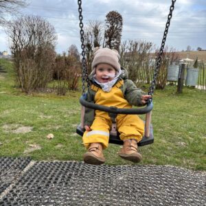 Ein kleiner Junge in gelber Kleidung schwingt fröhlich auf einer Schaukel im Freien und lächelt in die Kamera.