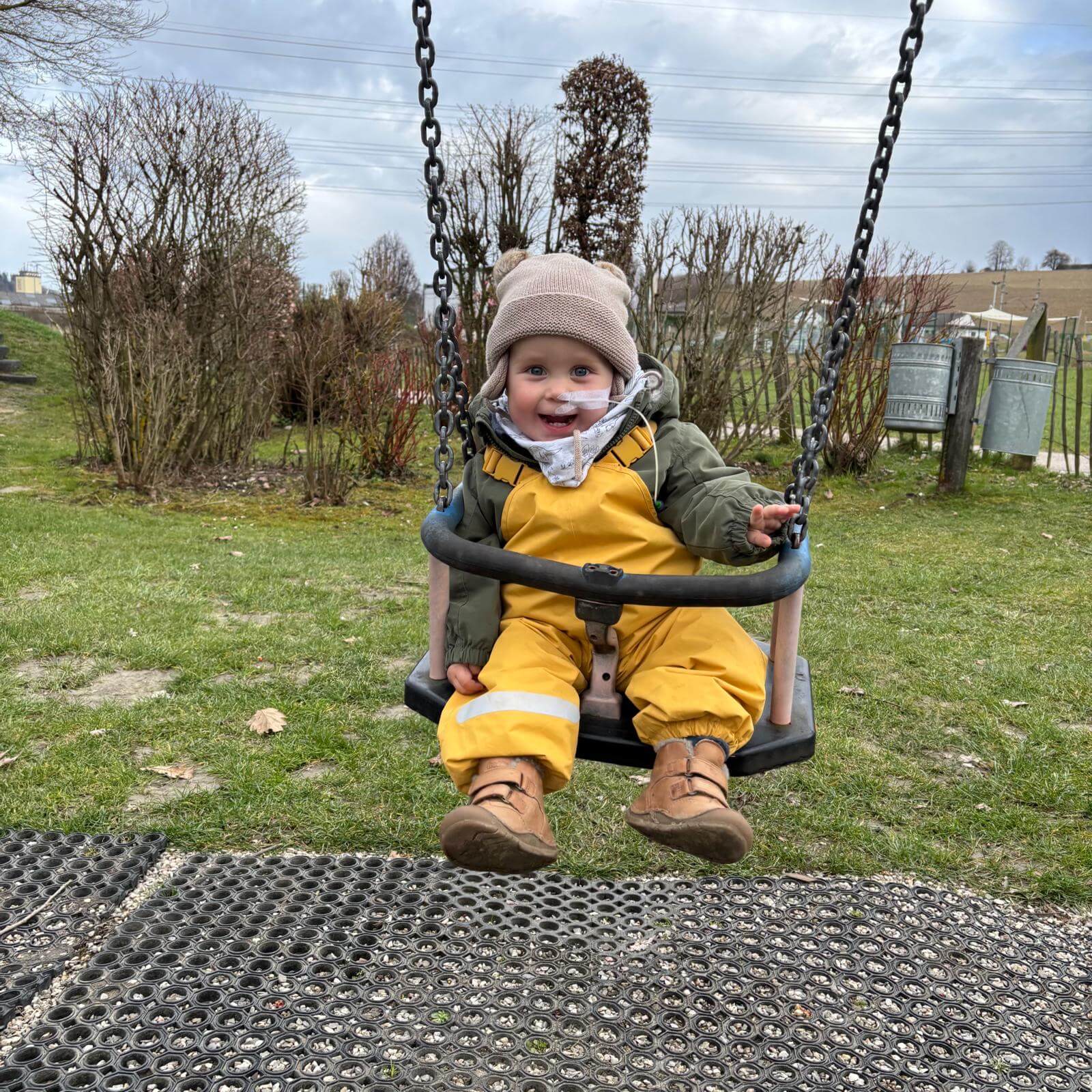 Ein kleiner Junge in gelber Kleidung schwingt fröhlich auf einer Schaukel im Freien und lächelt in die Kamera.