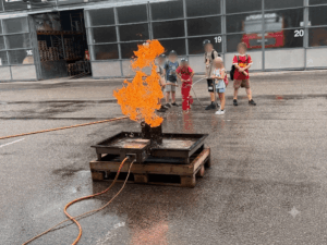 Eine Gruppe von Kindern beobachtet eine kontrollierte Feuerübung mit orangefarbenen Flammen in einer sicheren Umgebung.