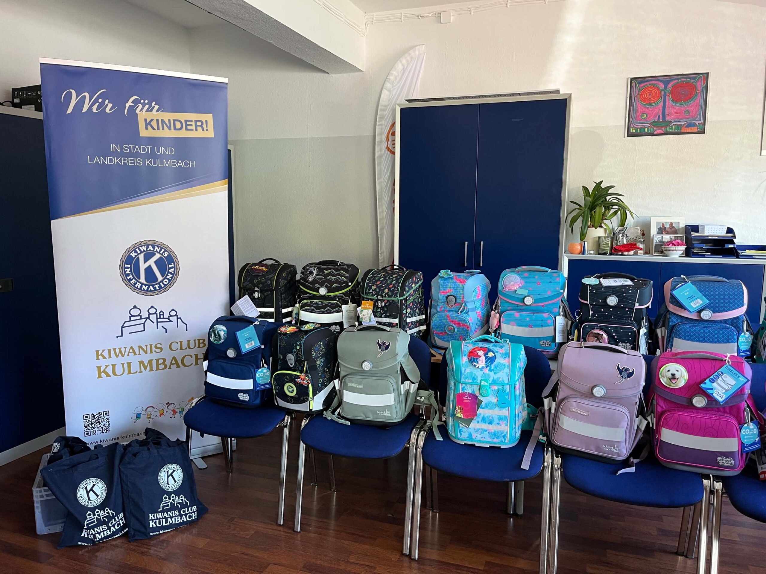 Diverse Schulranzen stehen auf blauen Stühlen, daneben ein Banner des Kiwanis Clubs Kulmbach mit der Aufschrift "Wir für Kinder!".