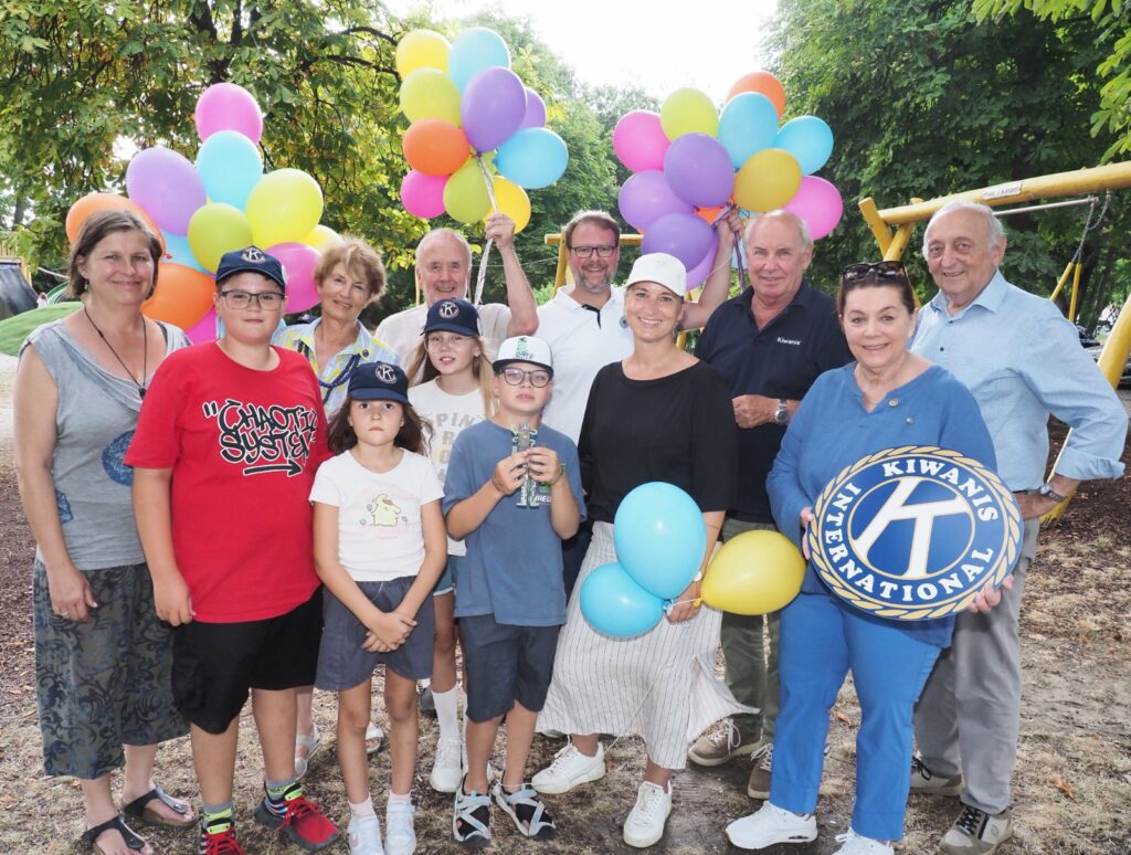 Eine Gruppe von Erwachsenen und Kindern hält bunte Luftballons und lächelt, während sie vor einem Spielplatz posiert.