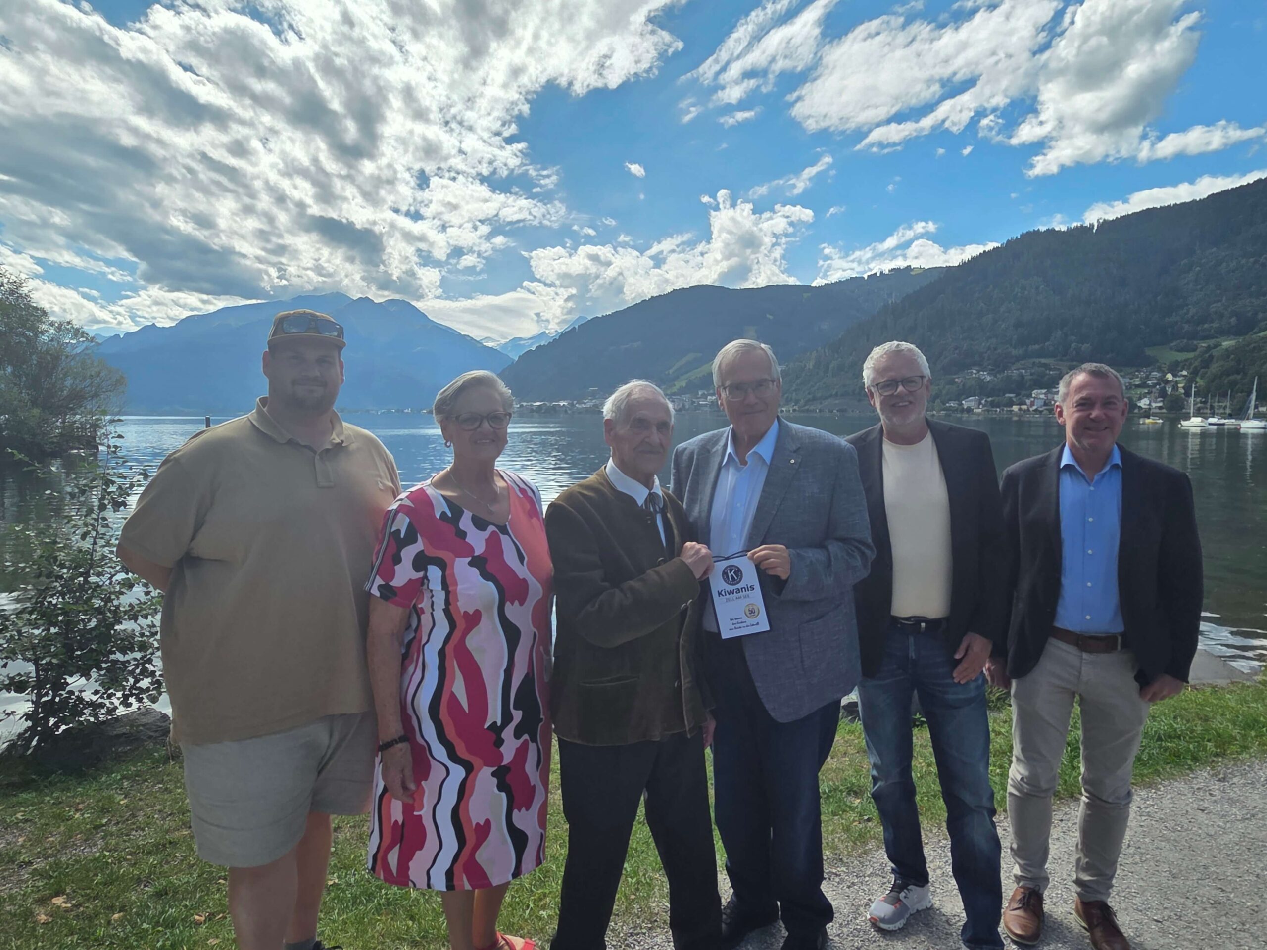 Sechs Personen stehen am Ufer eines Sees, im Hintergrund Berge und bewölkter Himmel, eine Person hält ein Schild in der Hand.