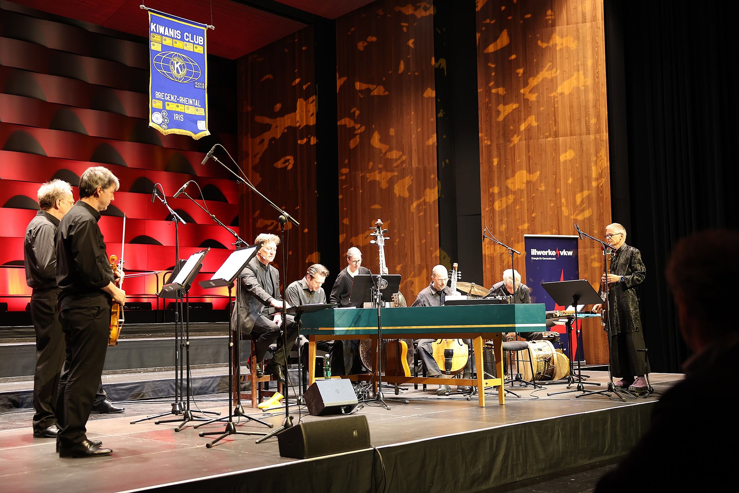Eine Gruppe von Musikern spielt auf der Bühne, umgeben von verschiedenen Instrumenten und einer farbigen Hintergrundbeleuchtung.