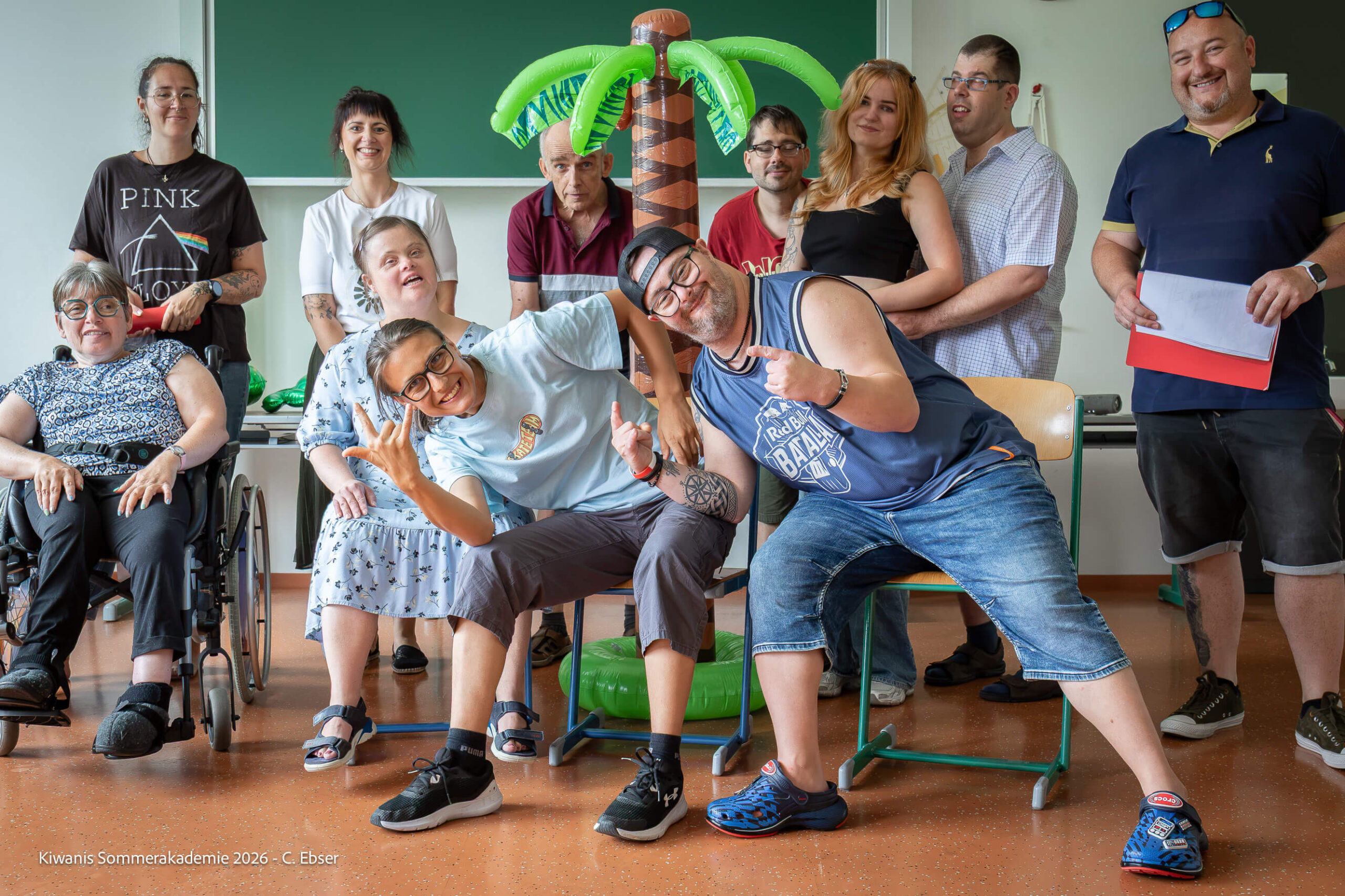 Eine Gruppe von Menschen unterschiedlichen Alters und mit Behinderungen posiert fröhlich vor einer aufblasbaren Palme in einem Klassenraum.