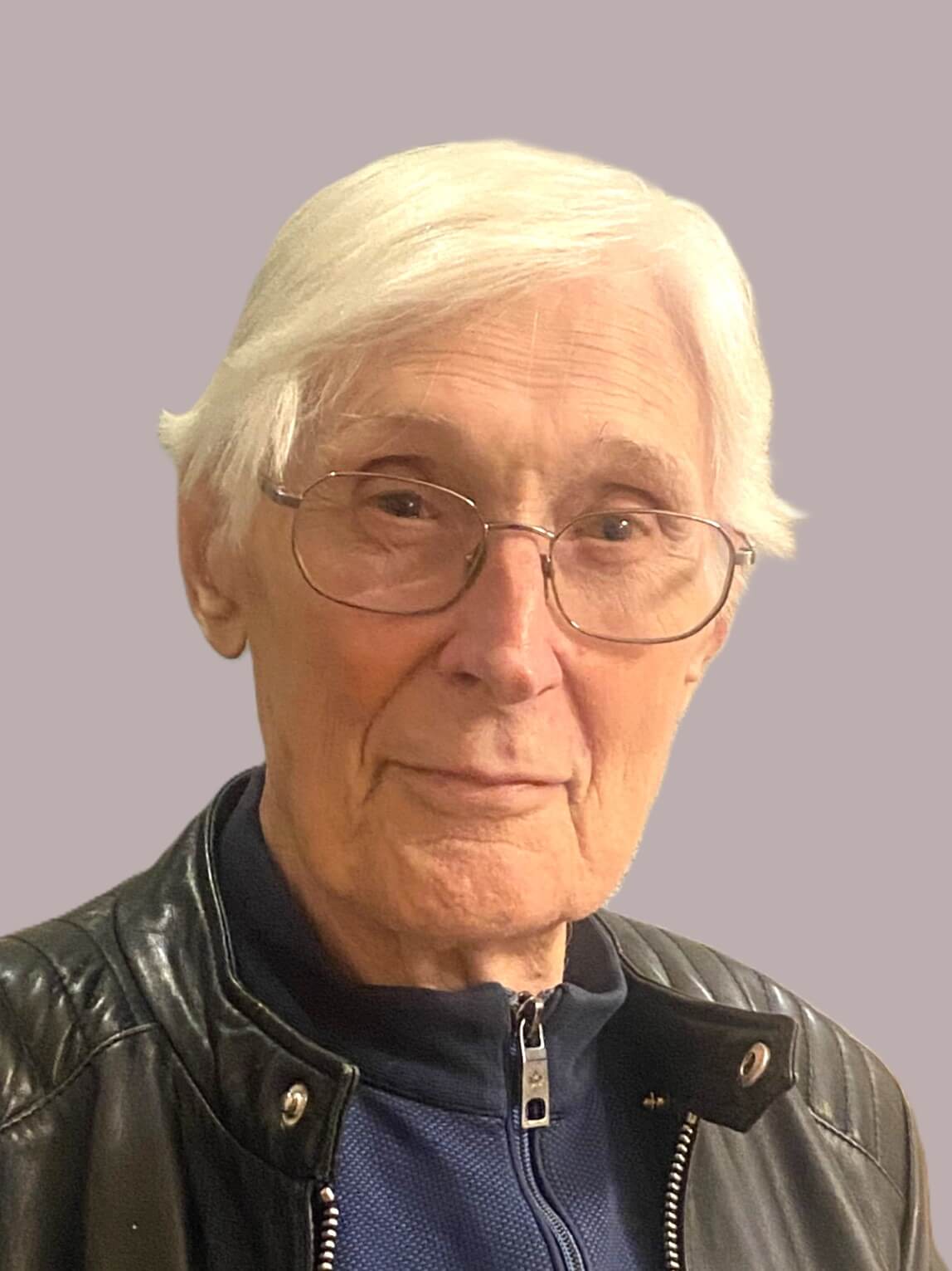Älterer Mann mit Brille und weißen Haaren, trägt eine schwarze Jacke, lächelt vor einem einfarbigen Hintergrund.