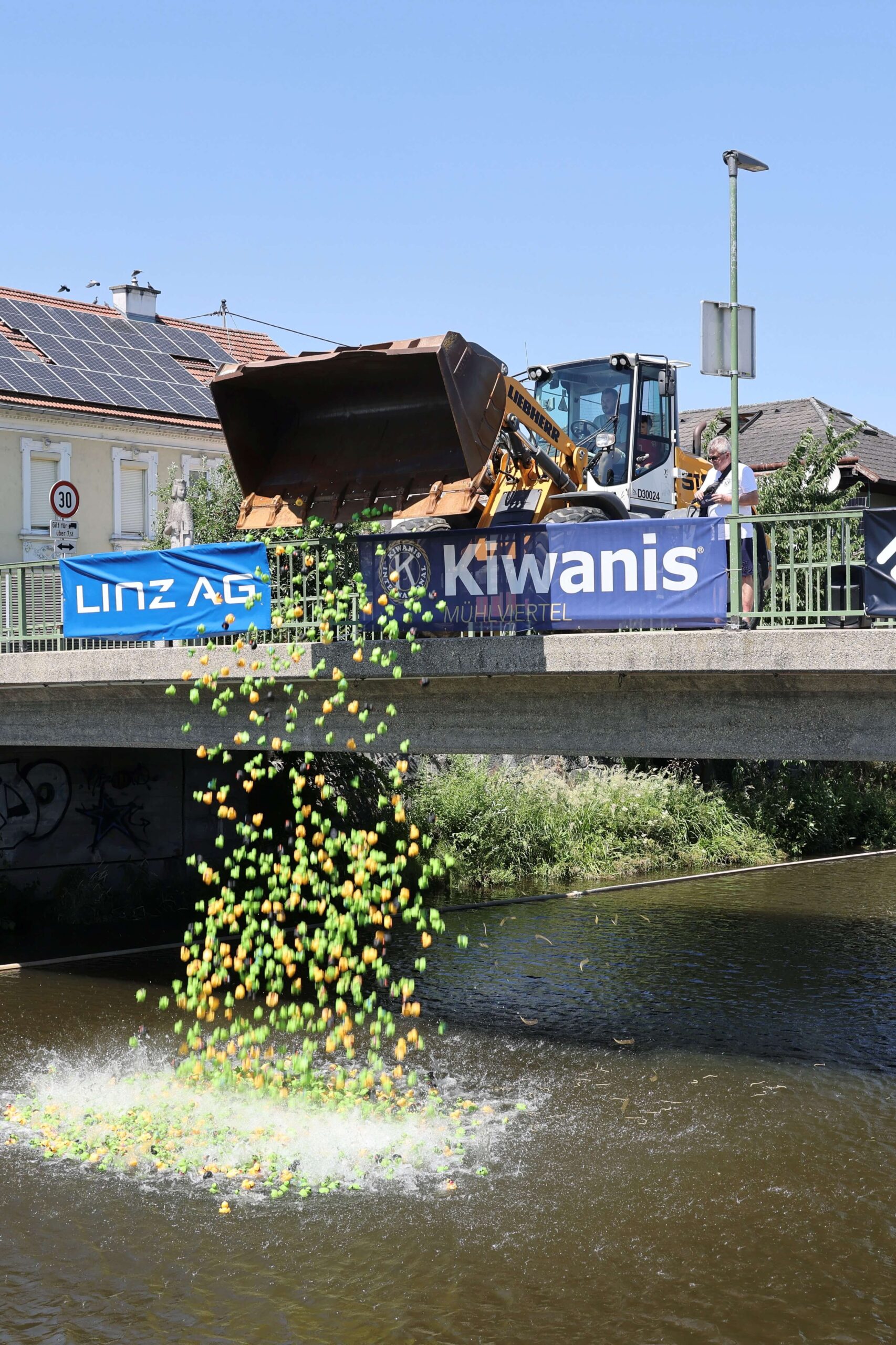 Ein Bagger kippt gelbe und grüne Enten in einen Fluss unter einer Brücke, während zwei Banner sichtbar sind.