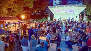 Belebte Freiluftveranstaltung mit vielen Menschen an Tischen, Bühne mit Musikern und festlicher Atmosphäre bei Nacht.