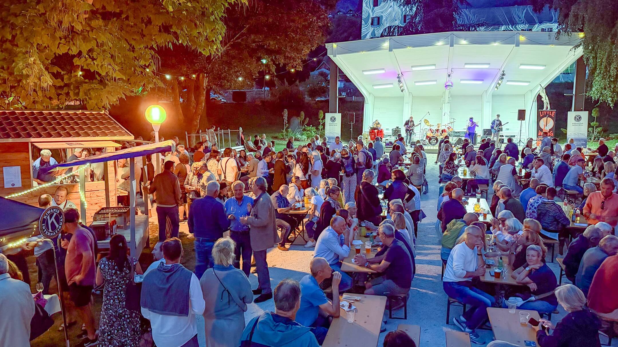Belebte Freiluftveranstaltung mit vielen Menschen an Tischen, Bühne mit Musikern und festlicher Atmosphäre bei Nacht.