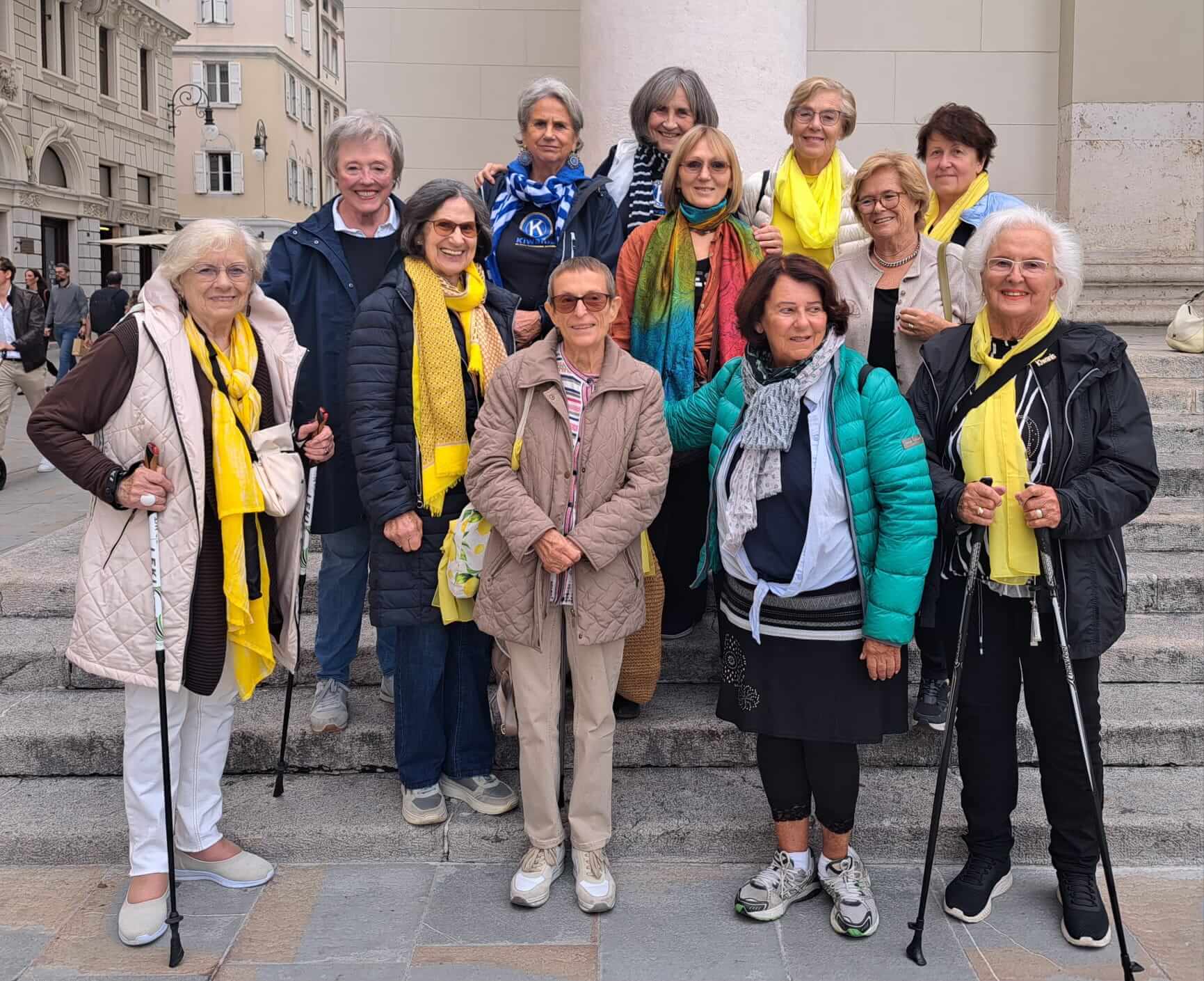 Eine Gruppe von zwölf Frauen in bunten Schals und Jacken posiert fröhlich auf einer Treppe in einer Stadt.