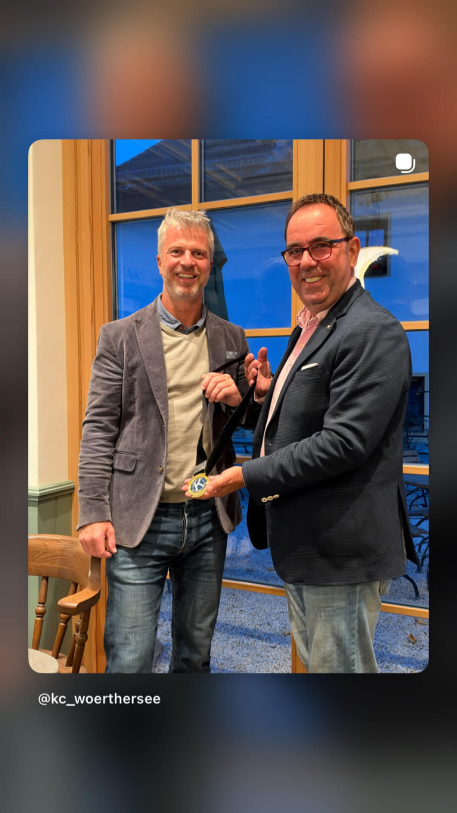 Zwei Männer stehen lächelnd in einem Raum und halten eine Medaille in der Hand; eine Fensterfront ist im Hintergrund sichtbar.