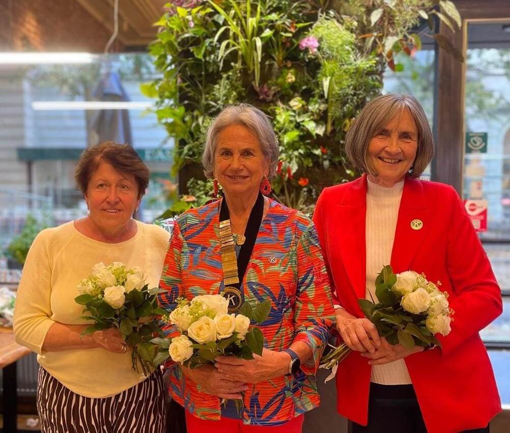 Drei Frauen mit Blumenarrangements stehen vor einer grünen Pflanzenwand in einem hellen Raum.