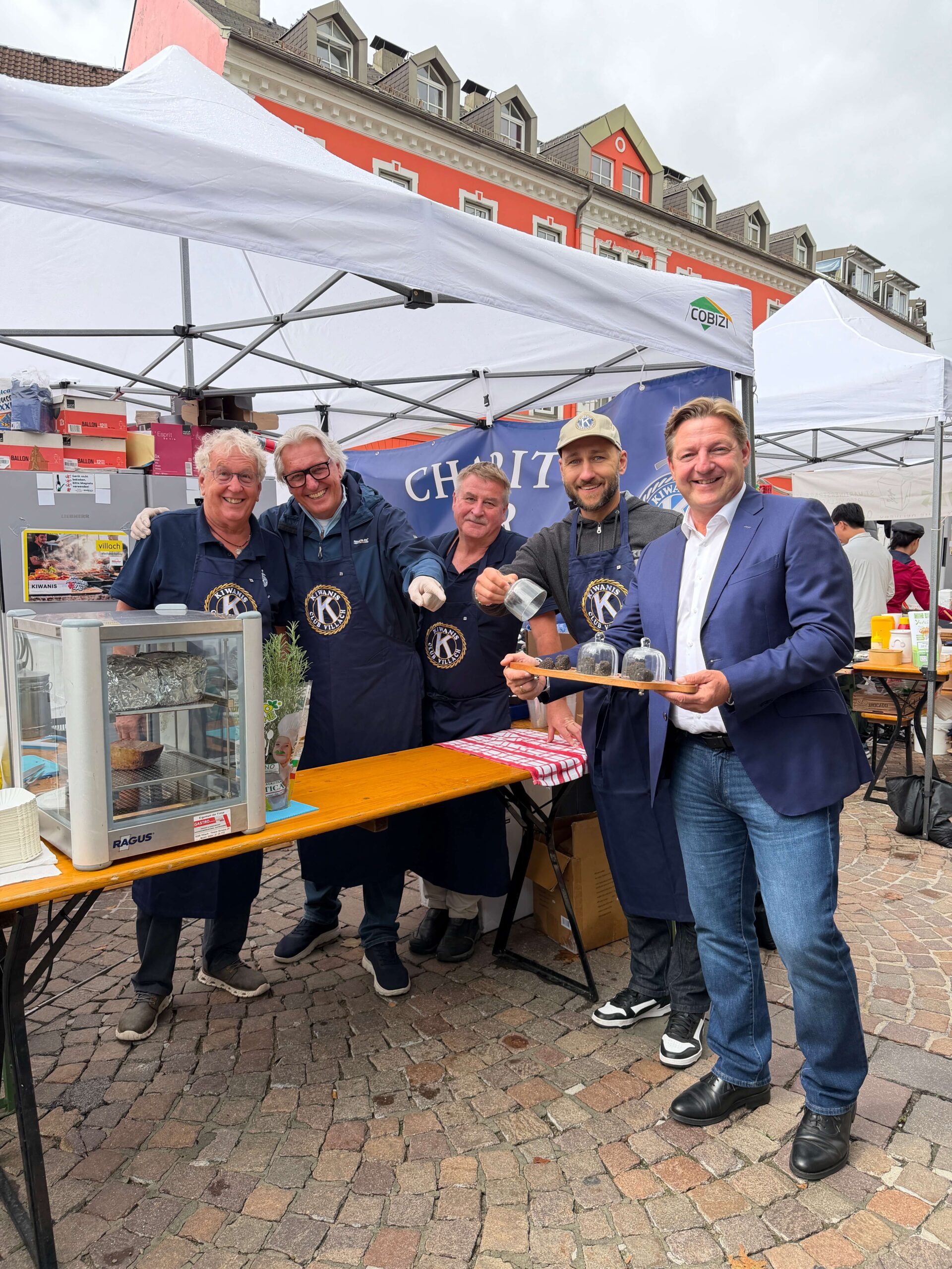 Sechs Männer in blauen Schürzen stehen an einem Marktstand und präsentieren Speisen vor einer Festzelt-Atmosphäre.