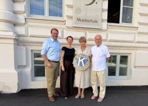 Vier Personen stehen vor einer Musikschule und halten ein Schild der Organisation Kiwanis International.