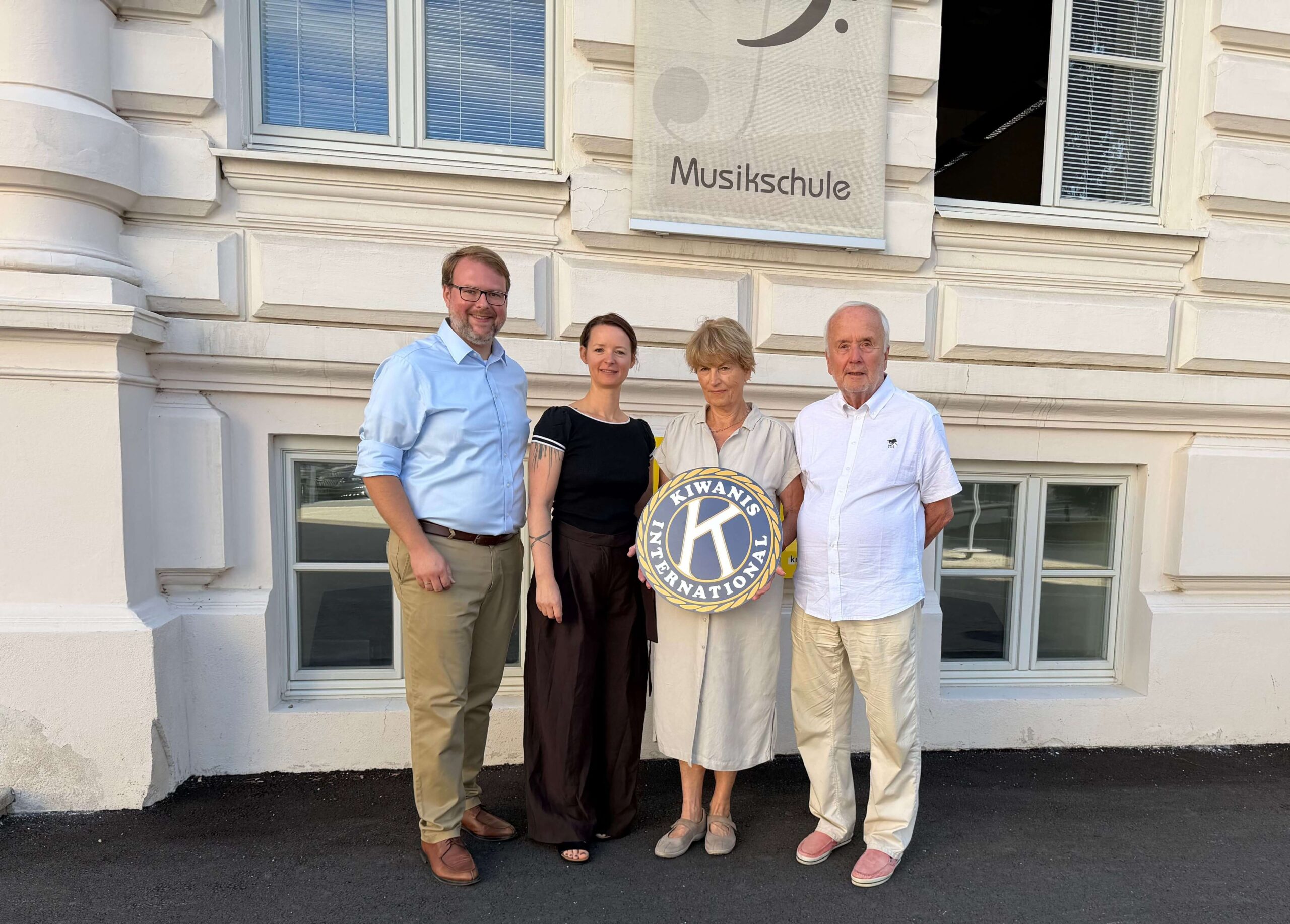 Vier Personen stehen vor einer Musikschule und halten ein Schild der Organisation Kiwanis International.