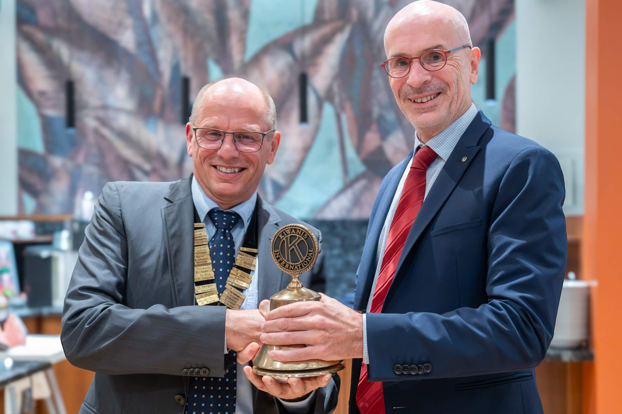 Zwei Männer in Anzügen halten gemeinsam eine goldene Glocke mit dem Schriftzug "Kiwanis International" in einer hellen Umgebung.