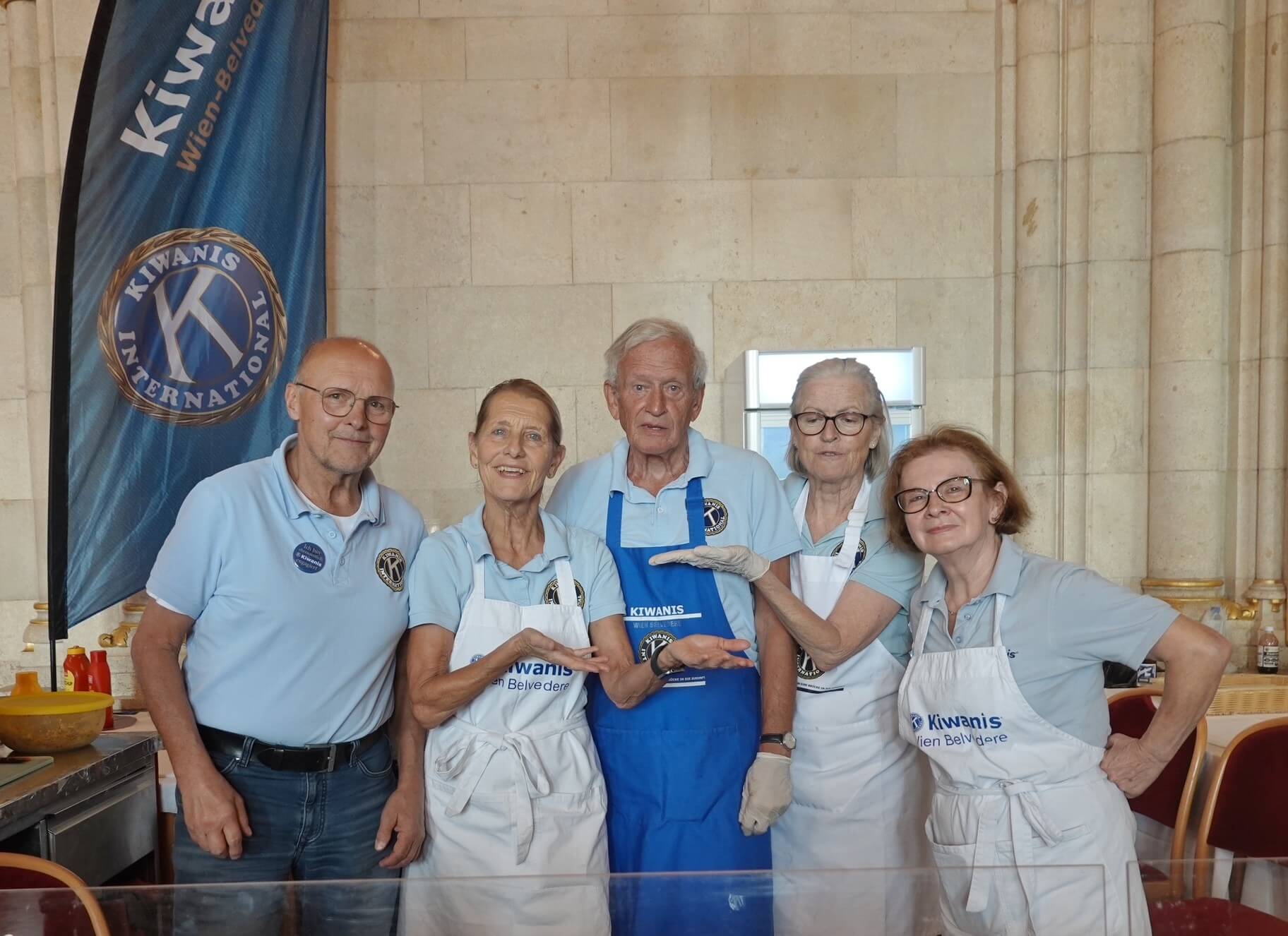Fünf Personen in blauen Shirts und Schürzen posieren freundlich vor einem Kiwanis-Stand in einem hellen Raum.