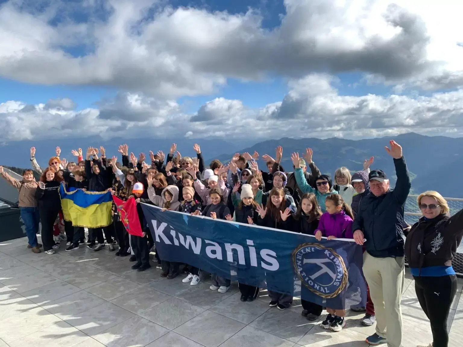 Eine große Gruppe von Menschen steht auf einem Berggipfel, winkt und hält ein Banner der Organisation Kiwanis und eine ukrainische Flagge.